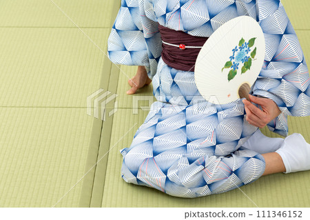 日式房間裡,一位穿著浴衣的中年婦女坐在榻榻米上,手裡拿著扇子 日式房間裡,一位穿著浴衣的中年婦女坐在榻榻米上,手裡拿著扇子 111346152