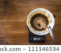 Pouring water on the light roast coffee grounds through the dripper with filter paper. On digital scale, placed on the wooden table. In top view. 111346594