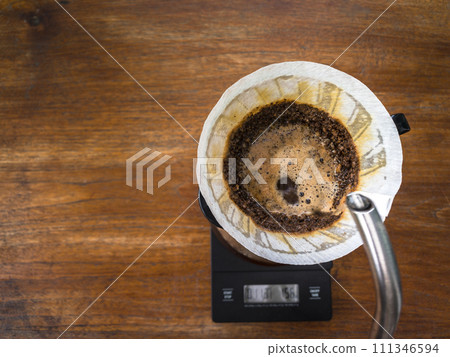 Pouring water on the light roast coffee grounds through the dripper with filter paper. On digital scale, placed on the wooden table. In top view. Pouring water on the light roast coffee grounds through the dripper with filter paper. On digital scale, placed on the wooden table. In top view. 111346594
