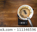 Pouring water on the light roast coffee grounds through the dripper with filter paper. On digital scale, placed on the wooden table. In top view. 111346596