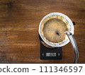 Pouring water on the light roast coffee grounds through the dripper with filter paper. On digital scale, placed on the wooden table. In top view. 111346597