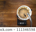 Pouring water on the light roast coffee grounds through the dripper with filter paper. On digital scale, placed on the wooden table. In top view. 111346598