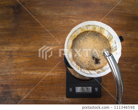 Pouring water on the light roast coffee grounds through the dripper with filter paper. On digital scale, placed on the wooden table. In top view. Pouring water on the light roast coffee grounds through the dripper with filter paper. On digital scale, placed on the wooden table. In top view. 111346598