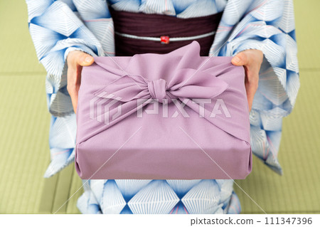 一位身穿浴衣的中年婦女坐在日式房間的榻榻米上，手裡拿著紫色的包袱皮。 111347396