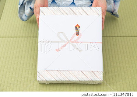 一位穿著浴衣的中年婦女坐在日式房間的榻榻米上,手裡拿著年中禮物 一位穿著浴衣的中年婦女坐在日式房間的榻榻米上,手裡拿著年中禮物 111347815