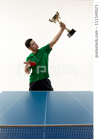 Player in victory pose holds cup and racquet at table tennis match against white studio background. Power and strength. Player in victory pose holds cup and racquet at table tennis match against white studio background. Power and strength. 111348021