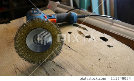 hand-held electric tool with a round brush with metallic golden bristles for processing wooden material, lying on a workbench in a carpentry workshop, a screwdriver with an emery attachment 111349934
