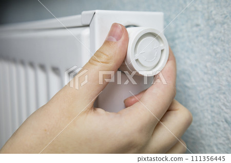 Woman regulates heating on white battery radiator at home in cold winter season, hands close-up. Central heating in well-maintained modern building. Adjustment sensor of heater to change temperature Woman regulates heating on white battery radiator at home in cold winter season, hands close-up. Central heating in well-maintained modern building. Adjustment sensor of heater to change temperature 111356445