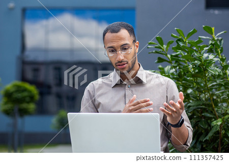 Handsome african american remote worker in eyewear sitting with laptop outdoors on bench by bush. Attentive project manager hosting video conference and explaining progress strategy for colleagues. 111357425