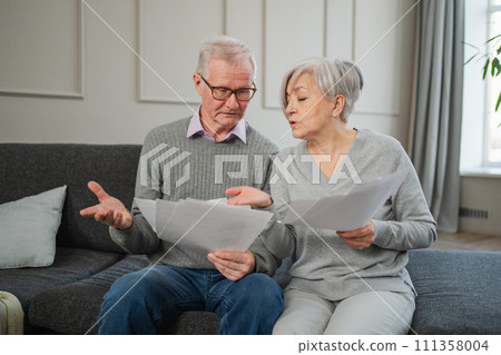 Sad tired disappointed middle aged senior couple sit with paper document. Unhappy older mature man woman reading paper bill managing bank finances calculating taxes planning loan debt pension payment 111358004