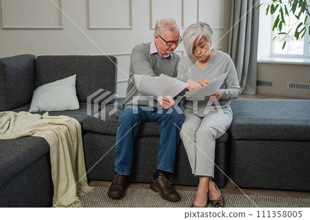Sad tired disappointed middle aged senior couple sit with paper document. Unhappy older mature man woman reading paper bill managing bank finances calculating taxes planning loan debt pension payment 111358005