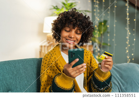 African american woman shopping online holding smartphone paying with gold credit card. Girl sitting at home buying on Internet enter credit card details. Online shopping ecommerce delivery service 111358551