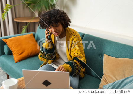 African American woman freelancer using laptop talking on phone at home office. Happy business girl having virtual meeting. Woman using phone laptop for communication working indoors. Remote work 111358559