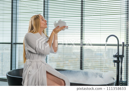 Spa relaxation. Young woman in spa bathrobe sitting at bath blowing foam in bathtub. Girl relaxing in bathroom at home. Female spending morning in bathroom. Beauty treatment body skin care concept Spa relaxation. Young woman in spa bathrobe sitting at bath blowing foam in bathtub. Girl relaxing in bathroom at home. Female spending morning in bathroom. Beauty treatment body skin care concept 111358633