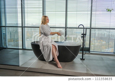 Spa relaxation. Young woman in spa bathrobe sitting at bath with foam in bathtub. Girl relaxing in bathroom at home. Female spending morning in bathroom. Beauty treatment body skin care concept 111358635