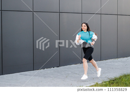 Young woman with fit body jumping and running against dark wall background. Female model in sportswear exercising outdoors. 111358829
