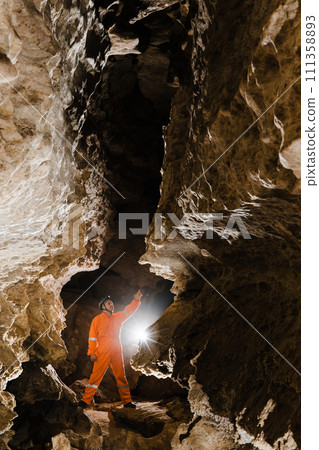 Man walking and exploring dark cave with light headlamp underground. Mysterious deep dark, explorer discovering mystery moody tunnel looking on rock wall inside. 111358893