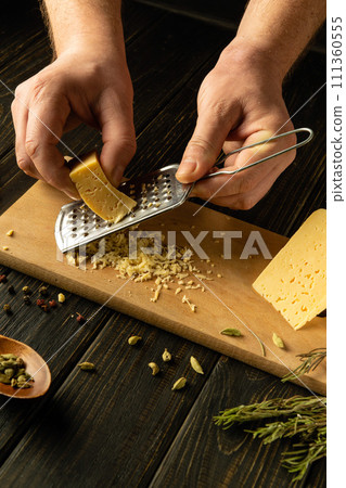 Grating hard cheese with a grater in the hand of a cook for preparing breakfast on the kitchen table. Cheese dish concept. 111360555