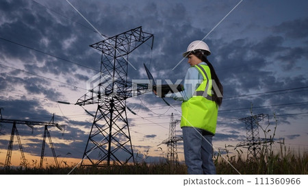 Woman electrician marks notes online on laptop observing power transmission lines at twilight countryside closeup. Engineer tests system with laptop at power generation plant under dramatic sky 111360966