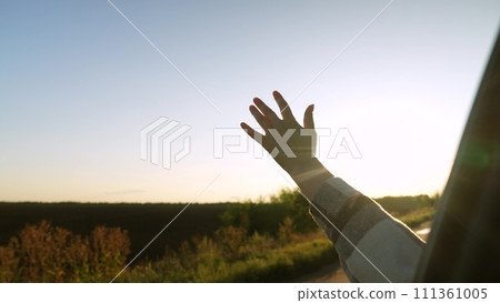 Road trip female hand from window of driving car at sunset sunrise rural summer field closeup. Woman arm enjoy freedom air travel adventure active lifestyle automobile journey exploration discovery 111361005