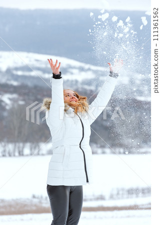 Full-length portrait of girl in white down jacket tossing snow. Winter smiling woman. Vertical frame. Full-length portrait of girl in white down jacket tossing snow. Winter smiling woman. Vertical frame. 111361562