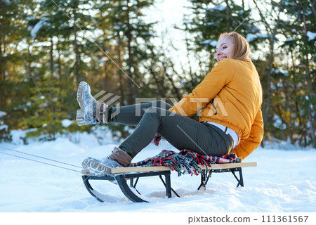 Young woman in yellow jacket rides sled through snow-covered pine forest. Winter holidays. Young woman in yellow jacket rides sled through snow-covered pine forest. Winter holidays. 111361567