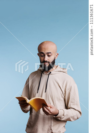 Arab man reading book paperback edition with yellow cover. Young concentrated bald bearded person in hoodie holding open notepad while standing with surprised facial expression Arab man reading book paperback edition with yellow cover. Young concentrated bald bearded person in hoodie holding open notepad while standing with surprised facial expression 111361671