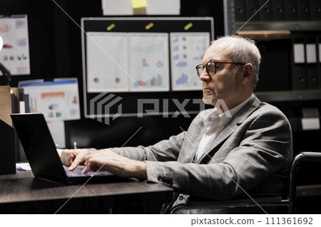 Senior accountant in wheelchair checking administrative analytical data in cabinet room office surrounded by accountancy statistic reports. Professional businessman in file storage room Senior accountant in wheelchair checking administrative analytical data in cabinet room office surrounded by accountancy statistic reports. Professional businessman in file storage room 111361692