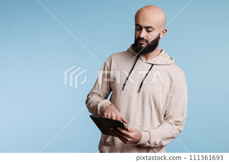 Arab man using digital tablet, surfing internet and tapping on touchscreen. Young focused person holding portable gadget, browsing social media networks and scrolling online pages Arab man using digital tablet, surfing internet and tapping on touchscreen. Young focused person holding portable gadget, browsing social media networks and scrolling online pages 111361693