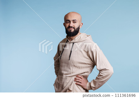 Arab man holding hands on waist while looking away with thoughtful facial expression. Young adult smiling person wearing casual hoodie clothes while standing in natural pose Arab man holding hands on waist while looking away with thoughtful facial expression. Young adult smiling person wearing casual hoodie clothes while standing in natural pose 111361696