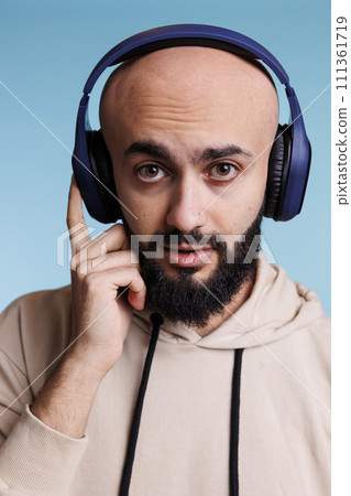 Young arab man listening to music in wireless headphones portrait. Bald bearded person enjoying tunes playlist in earphones while looking at camera with neutral expression 111361719