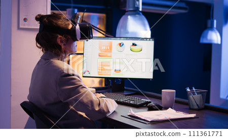 Corporate advisor planning report with vr glasses at desk, working with virtual reality 3d environment to increase profit at night. Young woman using 3d simulation goggles. Handheld shot. Corporate advisor planning report with vr glasses at desk, working with virtual reality 3d environment to increase profit at night. Young woman using 3d simulation goggles. Handheld shot. 111361771