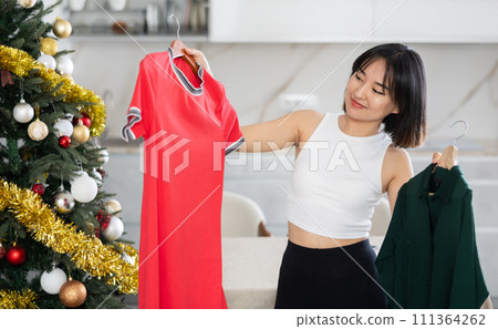 Happy young Asian girl trying to choose a dress standing next to Xmas-tree 111364262