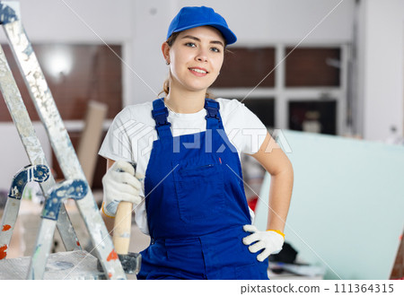 Portrait of a contented young female builder in blue overalls holding hammer Portrait of a contented young female builder in blue overalls holding hammer 111364315