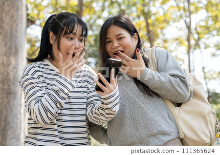 Two amazed young Asian female college students are surprised by unbelievable news on a smartphone. 111365624