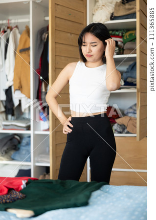 Sad young Asian girl trying to choose a dress standing by wardrobe Sad young Asian girl trying to choose a dress standing by wardrobe 111365885