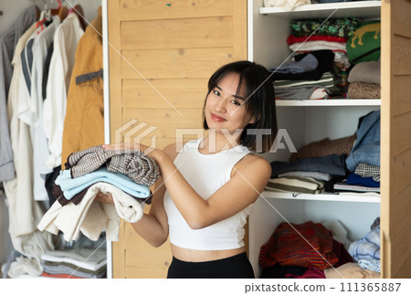 Happy young Asian girl holding some items of clothing by the wardrobe 111365887
