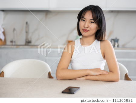 Smiling young Asian girl sitting in the kitchen with mobile phone on the table 111365981