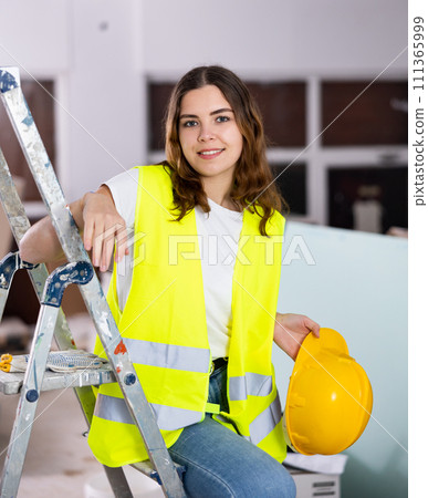 Portrait of young woman in protective yellow vest and hard hat in room being renovated 111365999