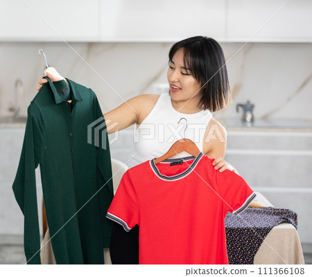 Happy young Asian girl trying to choose a dress in the kitchen Happy young Asian girl trying to choose a dress in the kitchen 111366108
