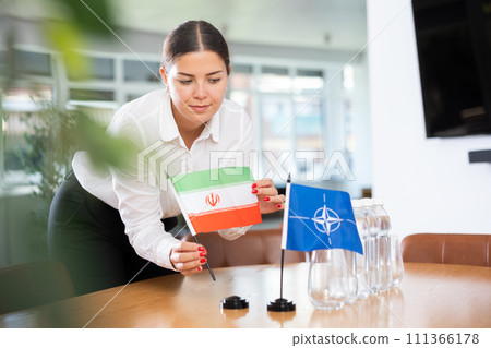 Woman secretary prepares an office for negotiations - she places flags of NATO (OTAN) and Iran table 111366178