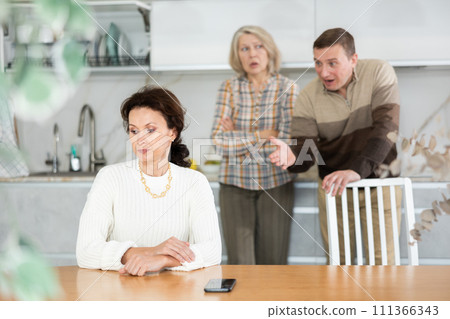 Angry old woman and middle-aged man quarreling to woman sitting at the kitchen table Angry old woman and middle-aged man quarreling to woman sitting at the kitchen table 111366343