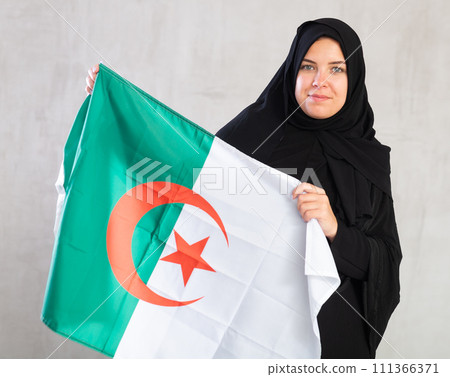 Balanced Muslim woman in traditional black hijab holds flag of Algeria Balanced Muslim woman in traditional black hijab holds flag of Algeria 111366371