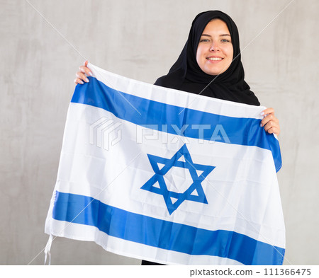 Balanced smiling Muslim woman in traditional black hijab holds flag of Israel Balanced smiling Muslim woman in traditional black hijab holds flag of Israel 111366475