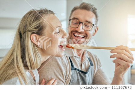 Happy couple cooking, tasting spoon of food and meal, lunch and dinner in kitchen at home. Smile mature man giving hungry woman a taste of homemade tomato sauce meal in mouth to enjoy eating together 111367673