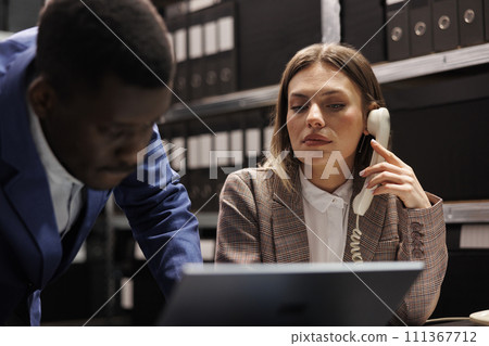 Secretary talking with bookkeeper at landline phone, working together at accountancy report in corporate depository. Diverse businesspeople analyzing administrative documents in file room 111367712