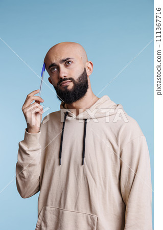 Pensive arab man holding pen to head while thinking and looking away. Young arabian person wearing casual hoodie making decision and remembering with thoughtful expression 111367716