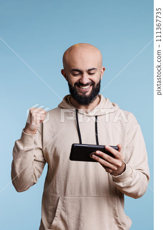 Excited arab man showing winner gesture with fist while playing online game on smartphone. Happy smiling gamer celebrating victory while entertaining with mobile phone videogame Excited arab man showing winner gesture with fist while playing online game on smartphone. Happy smiling gamer celebrating victory while entertaining with mobile phone videogame 111367735