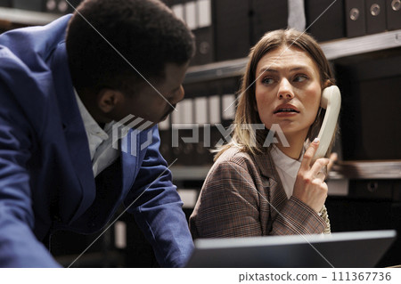 Caucasian detective using landline phone to talk with remote police officer, working at criminal case in arhive room. Diverse investigators analyzing crime scene evidence, trying to catch suspect Caucasian detective using landline phone to talk with remote police officer, working at criminal case in arhive room. Diverse investigators analyzing crime scene evidence, trying to catch suspect 111367736