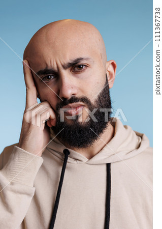 Arab man grimacing in pain while having migraine and holding head with finger portrait. Young bearded person rubbing temples while suffering from headache and looking at camera Arab man grimacing in pain while having migraine and holding head with finger portrait. Young bearded person rubbing temples while suffering from headache and looking at camera 111367738